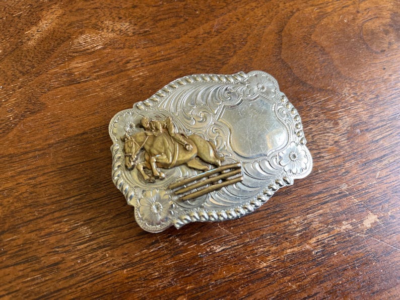May include: A rectangular silver-tone belt buckle with a rope-style border and intricate floral scrollwork. The center features a raised brass-colored relief of a horse and rider jumping over a fence, alongside a smooth, blank shield-shaped area.