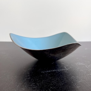 Puede incluir: Un cuenco decorativo con forma cuadrada y bordes curvos. El interior es de color azul claro, contrastando con el exterior oscuro. El cuenco es de metal y está sobre una superficie oscura.