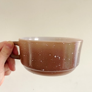 Puede incluir: Una taza de cerámica marrón y blanca moteada con asa. La taza tiene una forma ancha y redondeada y un interior blanco. El exterior es un degradado de marrón con motas blancas. La taza está sujeta por una mano.