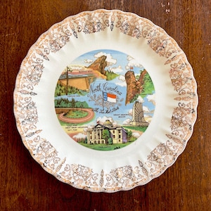 May include: A vintage decorative plate featuring a colorful illustration of North Carolina landmarks. The plate has a scalloped edge with a gold and white floral pattern. The center shows Blowing Rock, the Wright Memorial, and the State Capitol.
