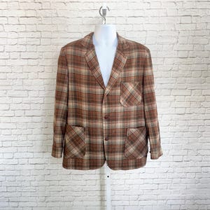 May include: A brown and tan plaid blazer featuring a notched lapel, two front buttons, one chest pocket, and two lower patch pockets. The garment is displayed on a mannequin, showcasing long sleeves and a structured fit with a classic collar design.