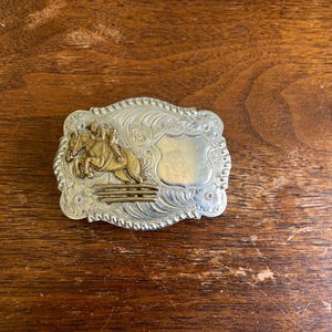 May include: A rectangular silver-toned belt buckle with a rope-style border and intricate floral scrollwork. The center features a raised gold-toned relief of a horse and rider jumping over a fence, alongside a smooth, blank shield-shaped plaque.