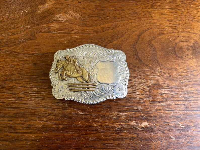 May include: A rectangular silver-toned belt buckle with a rope-style border and intricate floral scrollwork. The center features a raised gold-toned relief of a horse and rider jumping over a fence, alongside a smooth, blank shield-shaped plaque.