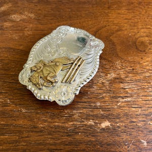 May include: A silver-toned belt buckle featuring an embossed floral scroll pattern and a central gold-toned relief of a horse and rider jumping over three parallel bars. The buckle has a scalloped edge and a smooth, polished shield-shaped area.