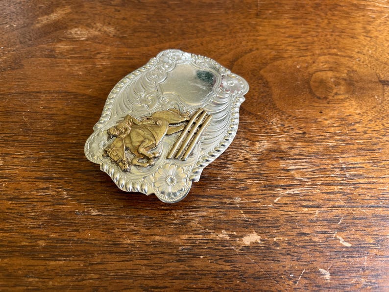 May include: A silver-toned belt buckle featuring an embossed floral scroll pattern and a central gold-toned relief of a horse and rider jumping over three parallel bars. The buckle has a scalloped edge and a smooth, polished shield-shaped area.