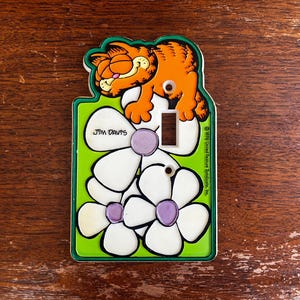 Puede incluir: Una cubierta de interruptor de luz vintage de Garfield. La cubierta presenta a Garfield el gato en naranja, descansando sobre flores blancas con centros morados. El fondo es verde con el texto "Jim Davis".
