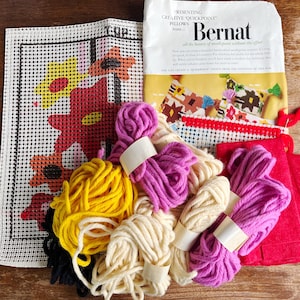 May include: A needlepoint craft kit featuring a white mesh canvas with a printed floral pattern, a Bernat Quickpoint instruction packet, and multiple skeins of yarn in yellow, purple, cream, and red, along with a red fabric piece.