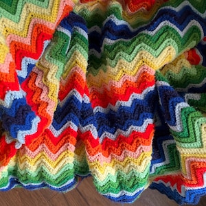 Puede incluir: Una manta de ganchillo hecha a mano con un vibrante patrón de chevrón en varios colores, incluyendo rojo, naranja, amarillo, verde claro, verde oscuro, azul claro y azul oscuro. El tejido muestra un diseño de punto en zigzag repetido.