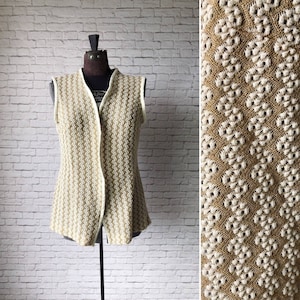 Könnte beinhalten: Eine ärmellose Vintage-Weste mit beigem und cremefarbenem Blumenmuster. Die Weste hat einen V-Ausschnitt und eine Knopfleiste vorne. Die cremefarbene Paspel umrandet den Halsausschnitt und die Armausschnitte. Die Weste wird auf einer Schaufensterpuppe präsentiert.