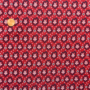 Könnte beinhalten: Ein roter Stoff mit einem sich wiederholenden Muster aus kleinen weißen und dunkelblauen blumenartigen Formen und winzigen Punkten. Eine kupferfarbene Münze liegt auf dem Stoff, um die Größe des Druckmusters zu verdeutlichen.