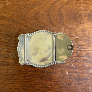 May include: A brass belt buckle featuring a rectangular central plate with a beaded border, a hinged metal bar for attachment, and a circular hook mechanism on the right side. The metal surface shows a smooth, polished finish with minor patina.