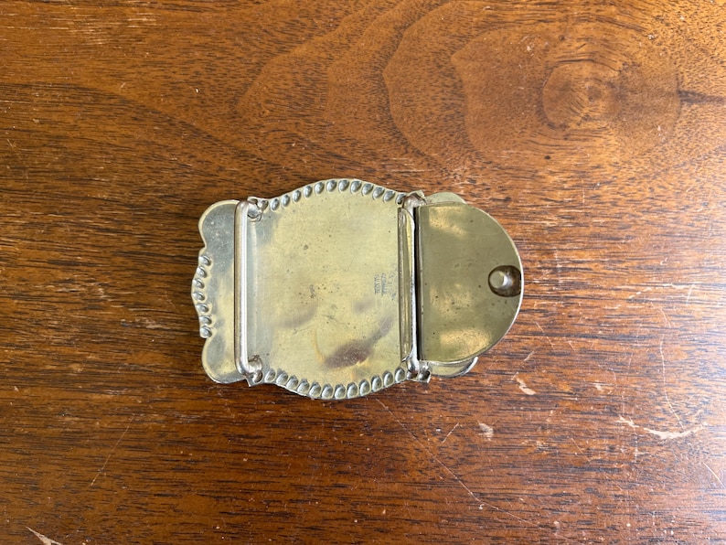 May include: A brass belt buckle featuring a rectangular central plate with a beaded border, a hinged metal bar for attachment, and a circular hook mechanism on the right side. The metal surface shows a smooth, polished finish with minor patina.