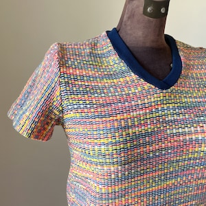 May include: A short-sleeved shirt featuring a vibrant, multi-colored woven pattern of red, yellow, blue, and white threads. The garment has a solid dark blue ribbed V-neck collar and a textured, grid-like appearance across the entire fabric surface.