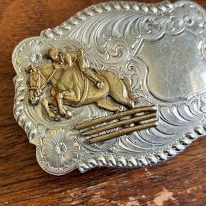 May include: A rectangular silver-tone belt buckle featuring an embossed floral scroll pattern and a rope-style border. The center displays a gold-tone relief of a rider on a horse jumping over a three-bar fence, alongside a smooth shield area.