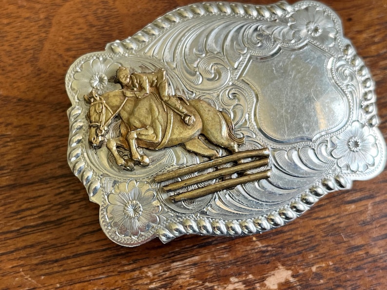 May include: A rectangular silver-tone belt buckle featuring an embossed floral scroll pattern and a rope-style border. The center displays a gold-tone relief of a rider on a horse jumping over a three-bar fence, alongside a smooth shield area.