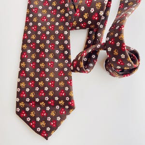 May include: A brown tie featuring a repeating pattern of small red, white, and gold flowers. The tie is knotted and rests on a white surface. The floral design is evenly distributed across the fabric.
