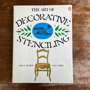 May include: A vintage book titled "The Art of Decorative Stenciling" with a white cover. The cover features a blue bird in a circle, green leaves, and a green chair with floral details. Authors are Adele Bishop and Cile Lord.