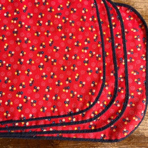 May include: A set of three red fabric placemats featuring a repeating pattern of small yellow and blue flowers interspersed with tiny white dots. Each placemat has rounded corners and is finished with a dark navy blue fabric binding edge.