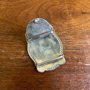 May include: A silver-toned metal belt buckle with a scalloped edge and a smooth, reflective surface. The back of the buckle features a hinged pin mechanism and a rectangular bar for attaching to a belt strap, showing a metallic finish.