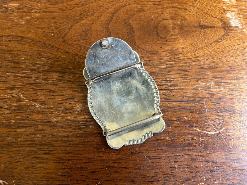 May include: A silver-toned metal belt buckle with a scalloped edge and a smooth, reflective surface. The back of the buckle features a hinged pin mechanism and a rectangular bar for attaching to a belt strap, showing a metallic finish.