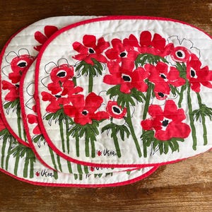 Puede incluir: Tres manteles individuales ovalados acolchados con fondo blanco y un vibrante estampado floral rojo con tallos verdes. Cada mantel tiene un borde de tela rojo brillante y el logotipo de Vera con un pequeño icono de mariquita impreso.