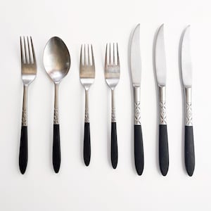 May include: A set of vintage silverware, including forks, spoons, and knives. The utensils have silver-colored metal heads and black handles. The handles feature a decorative pattern. The set is arranged on a white surface.