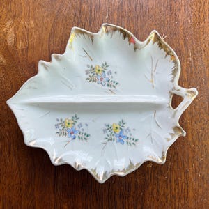 May include: A white, leaf-shaped divided dish with gold trim and floral accents. The dish is divided into two sections and features delicate blue, yellow, and pink flowers. The dish is likely vintage and is set on a wooden surface.