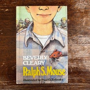 Puede incluir: Portada del libro "Ralph S. Mouse" de Beverly Cleary, ilustrado por Paul O. Zelinsky. La ilustración muestra a un niño con un ratón en el bolsillo. El título es amarillo.