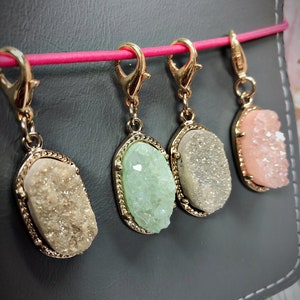 May include: Four gemstone charms in gold-colored settings hang from a pink cord. The gemstones are in shades of beige, green, gray, and pink. The charms are oval-shaped and have a textured, crystal-like appearance.
