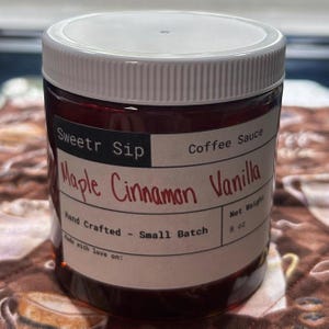 Peut inclure: Un pot de sauce au café "Sweetr Sip" avec un couvercle blanc. L'étiquette indique "Maple Cinnamon Vanilla" et "Hand Crafted - Small Batch". Le poids net est de 227g. Le pot est rempli d'un liquide brun foncé.