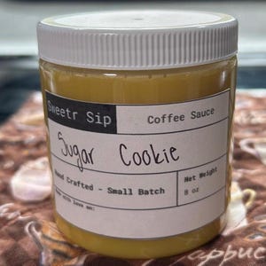 Può includere: Un barattolo di plastica trasparente con coperchio bianco riempito con un liquido color oro. L'etichetta recita "Sweetr Sip Coffee Sauce Sugar Cookie". Il barattolo è etichettato "Hand Crafted - Small Batch" e ha un peso netto di 227 grammi.