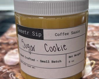 May include: A clear plastic jar with a white lid filled with a golden-colored liquid. The label reads "Sweetr Sip Coffee Sauce Sugar Cookie." The jar is labeled "Hand Crafted - Small Batch" and has a net weight of 8 oz.