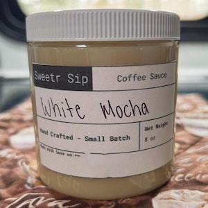 May include: A clear plastic jar with a white lid, filled with a light brown liquid. The label reads "Sweetr Sip Coffee Sauce White Mocha, Hand Crafted - Small Batch, Net Weight 227g."
