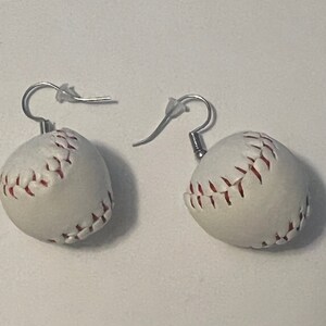 May include: A pair of baseball earrings. Each earring features a white baseball with red stitching and a silver-colored hook. The earrings are designed to resemble miniature baseballs.