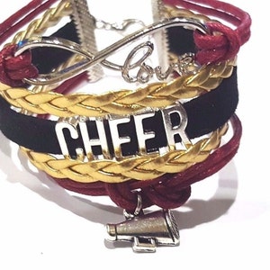 Può includere: Un braccialetto multistrato con cordini intrecciati rossi e dorati, una fascia in pelle scamosciata nera con la parola "CHEER" in lettere argentate e un simbolo di infinito argentato con la parola "Love" incisa. Un ciondolo a forma di megafono argentato pende dalla parte inferiore del braccialetto.