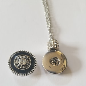 May include: A silver-toned necklace with a round pendant featuring a snap-on design. The pendant has a clear crystal center and a decorative black border. The necklace chain is delicate and the snap-on pendant is separate from the chain.