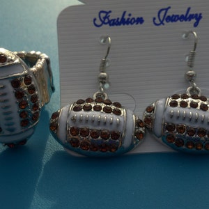 May include: A ring and a pair of earrings featuring a football design. The jewelry is white with brown accents and rhinestones. The earrings hang from silver hooks. The ring is adjustable. The jewelry is displayed on a Fashion Jewelry tag.