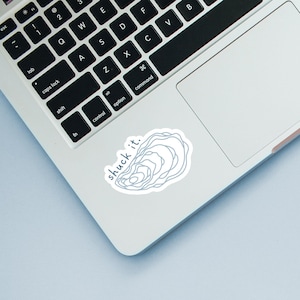 May include: A white sticker with a light blue outline of an oyster and the words "shuck it." The sticker is placed on a laptop, near the keyboard.