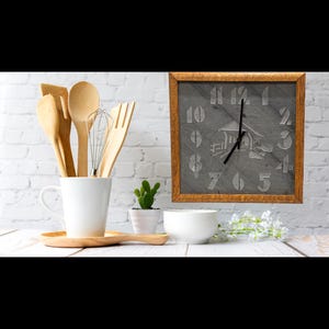 May include: A kitchen-themed still life featuring a wooden-framed clock with a house illustration, a white mug with wooden utensils, a small cactus, and a white bowl. The clock has a gray face with white numerals.