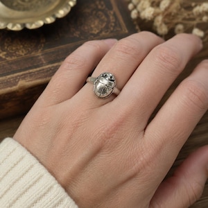 May include: A silver-toned ring featuring a detailed scarab beetle design on the face. The metal has an oxidized finish that highlights the textured lines and shape of the beetle. The band is simple and smooth, encircling the finger securely.