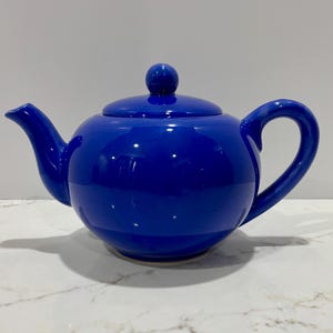 May include: A cobalt blue ceramic teapot with a matching lid and a rounded handle. The teapot has a glossy finish and is designed for brewing tea. The spout is curved, and the lid has a small knob for easy removal.