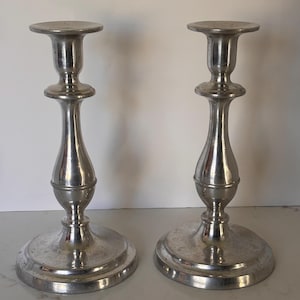 May include: Two silver-colored candlesticks with a classic design. Each candlestick has a round base, a slender stem, and a circular top to hold a candle. The candlesticks are made of metal and have a reflective surface.