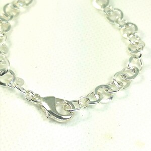 Op de afbeelding: Een zilverkleurige armband met een karabijnsluiting. De armband bestaat uit onderling verbonden, cirkelvormige schakels. De sluiting is glanzend, metaalachtig zilver. De armband wordt getoond tegen een witte achtergrond.
