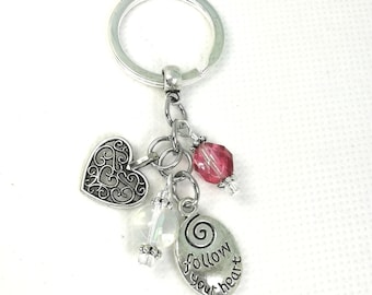 May include: Silver-toned keychain with multiple charms. Charms include a filigree heart, a clear crystal bead, a faceted pink bead, and an oval charm with the words "follow your heart".