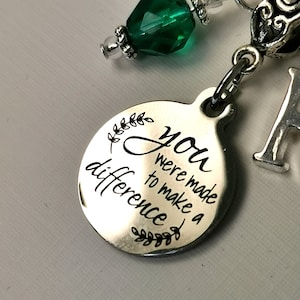 以下が含まれることがあります： 「You were made to make a difference」と黒い文字で書かれたシルバーカラーのペンダント。丸いペンダントには葉のデザインが施されています。緑色のファセットビーズとシルバーのイニシャルチャームも確認できます。
