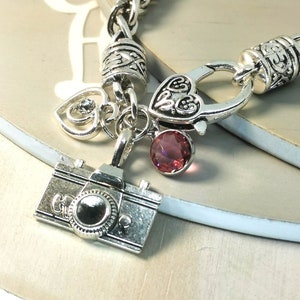 May include: Silver-toned charm bracelet featuring a camera charm, two heart charms, and a round, pink gemstone. The bracelet has a chain-link design and a clasp closure. The charms are detailed and the bracelet is displayed on a light-colored surface.