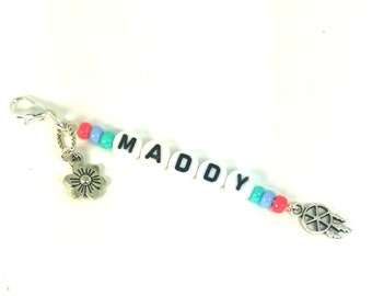 May include: A white beaded keychain with the name "MADDY" spelled out in black letters. The keychain has a silver flower charm on one end and a silver dreamcatcher charm on the other end.