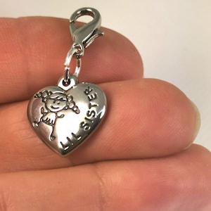 May include: Silver charm in the shape of a heart with a small cartoon girl and the words "Lil Sister" engraved on it. The charm is attached to a silver lobster clasp.