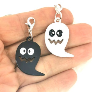 Op de afbeelding: Twee spookvormige bedels, een zwarte en een witte, met een zilveren sluiting. Elk spook heeft een glimlachend gezicht met twee ogen.