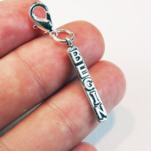 Op de afbeelding: Zilveren bedel met een zilveren clip. De bedel is een rechthoekige staaf met het woord "BEGIN" erop gegraveerd.